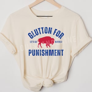 May include: Cream-colored t-shirt with the text "Glutton for Punishment" in blue, with a red buffalo graphic. The words "Let's Go Buffalo" are also included in the design.