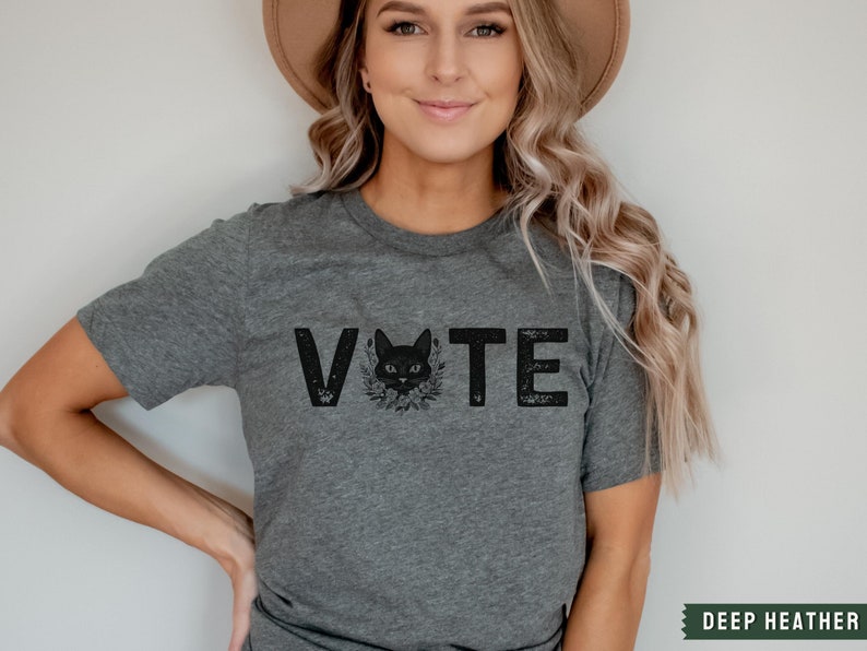 Puede incluir: Una camiseta gris con un gr&aacute;fico de un gato negro y la palabra "VOTE" impresa en ella. El gato lleva una corona de flores alrededor del cuello.