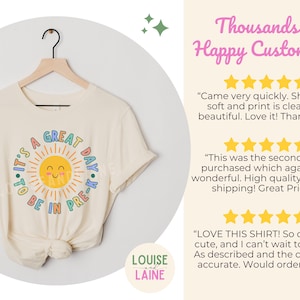 Peut inclure: Un t-shirt &agrave; manches courtes de couleur cr&egrave;me avec un motif de soleil jaune souriant entour&eacute; du texte It's a Great Day to be in Pre-K en lettres bleues, roses et orange. Le t-shirt est pr&eacute;sent&eacute; sur un cintre en bois sur un fond clair.