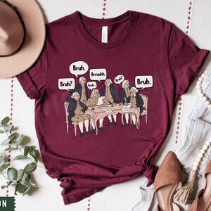 Peut inclure: Un t-shirt bordeaux avec un graphique des P&egrave;res fondateurs assis autour d'une table avec des bulles de dialogue qui disent "Bruh."