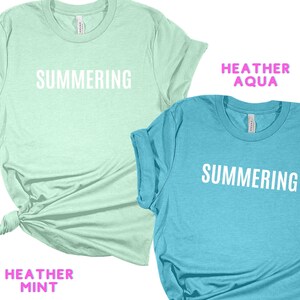 May include: Two t-shirts with the word "SUMMERING" printed on them. The shirt on the left is a light green color with the text "HEATHER MINT" below the design. The shirt on the right is a light blue color with the text "HEATHER AQUA" above the design.