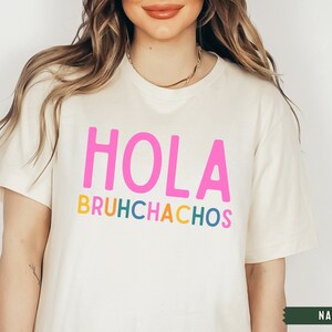 May include: A white t-shirt with the text "HOLA BRUHCHACHOS" in colorful letters. The text is in a playful font and the shirt is a casual style.