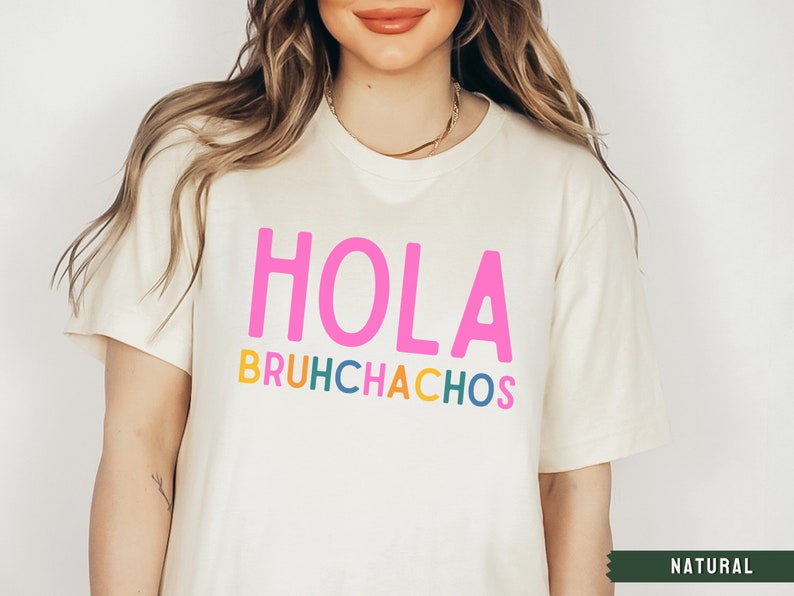 May include: A white t-shirt with the text "HOLA BRUHCHACHOS" in colorful letters. The text is in a playful font and the shirt is a casual style.