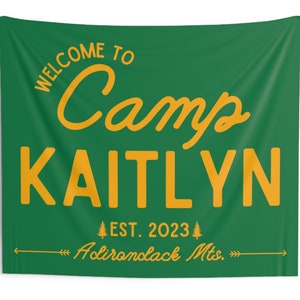 May include: A green and gold welcome to camp sign with the name Kaitlyn and the text "EST. 2023 Adirondack Mts." The sign is 80 inches by 68 inches.