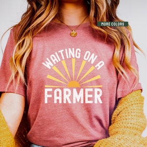 Może przedstawiać: R&oacute;żowy t-shirt z napisem "Waiting on a Farmer" w kolorze białym i ż&oacute;łtym graficznym słońcem.