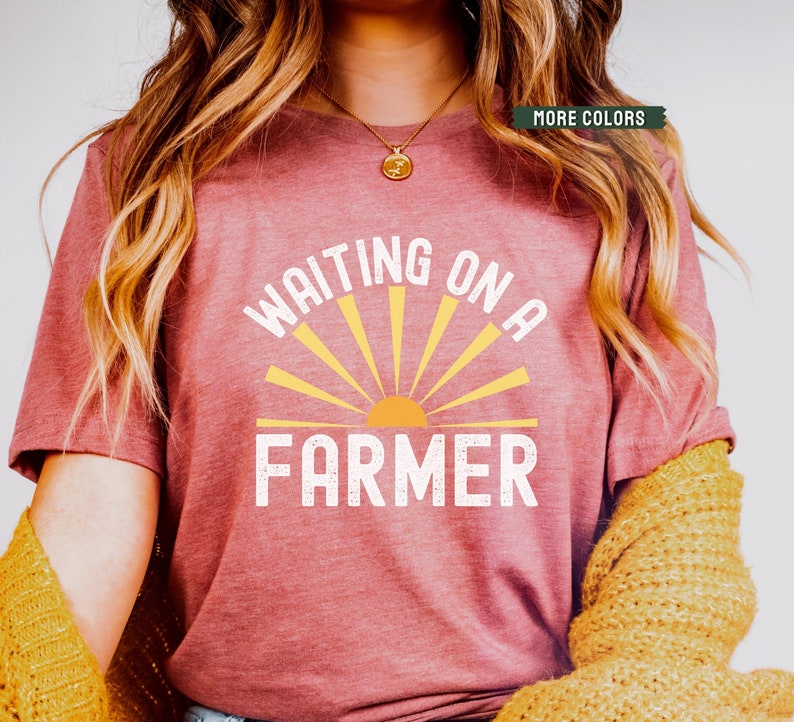 Może przedstawiać: R&oacute;żowy t-shirt z napisem "Waiting on a Farmer" w kolorze białym i ż&oacute;łtym graficznym słońcem.