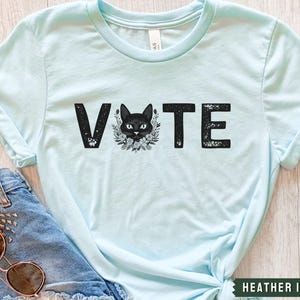 Puede incluir: Camiseta azul claro con un gr&aacute;fico de un gato negro y la palabra "VOTE" en letras negras desgastadas.