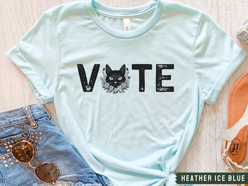 Puede incluir: Camiseta azul claro con un gr&aacute;fico de un gato negro y la palabra "VOTE" en letras negras desgastadas.