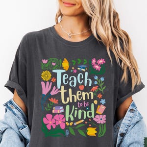 May include: A dark gray t-shirt with a colorful graphic design featuring flowers, books, pencils, and the text "Teach them to be kind".