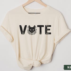 Puede incluir: Una camiseta de color crema con la palabra "VOTE" impresa en negro, con una silueta de gato negro en el medio de la palabra.