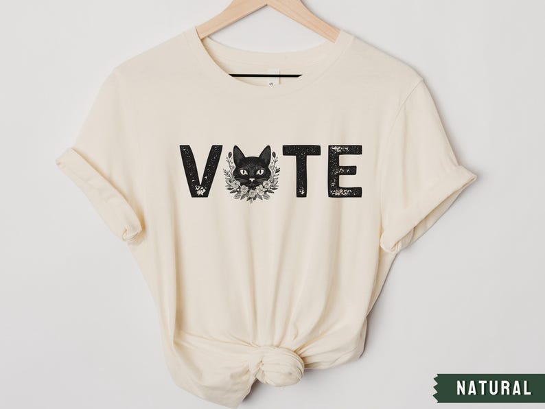 Puede incluir: Una camiseta de color crema con la palabra "VOTE" impresa en negro, con una silueta de gato negro en el medio de la palabra.