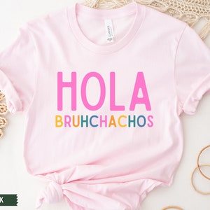 May include: A soft pink t-shirt with the text "HOLA BRUHCHACHOS" in colorful letters. The text is in a playful, casual font.