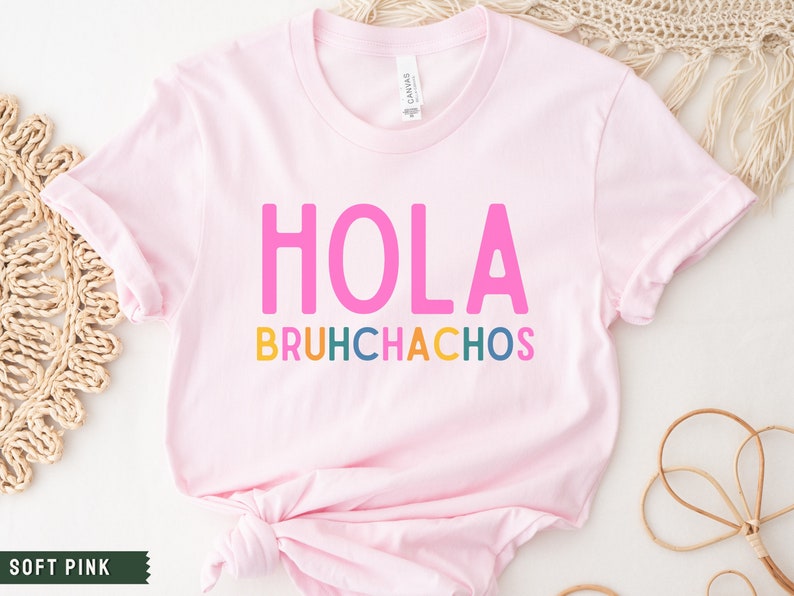 May include: A soft pink t-shirt with the text "HOLA BRUHCHACHOS" in colorful letters. The text is in a playful, casual font.