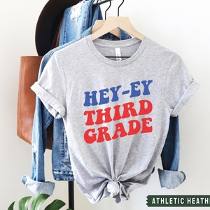 May include: A gray t-shirt with the text "HEY-EY THIRD GRADE" in red and blue letters. The shirt is on a hanger and has a knotted bottom. The text "ATHLETIC HEATHER" is visible at the bottom of the image.