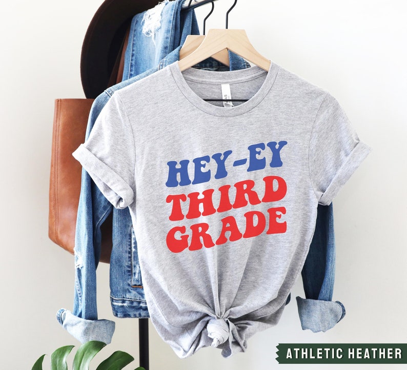 May include: A gray t-shirt with the text "HEY-EY THIRD GRADE" in red and blue letters. The shirt is on a hanger and has a knotted bottom. The text "ATHLETIC HEATHER" is visible at the bottom of the image.