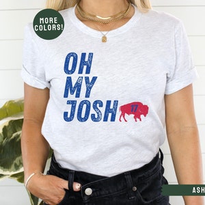 May include: A white t-shirt with blue text that reads "Oh My Josh" and a red and blue buffalo with the number 17 on it. The shirt is being worn by a person with a gold necklace.