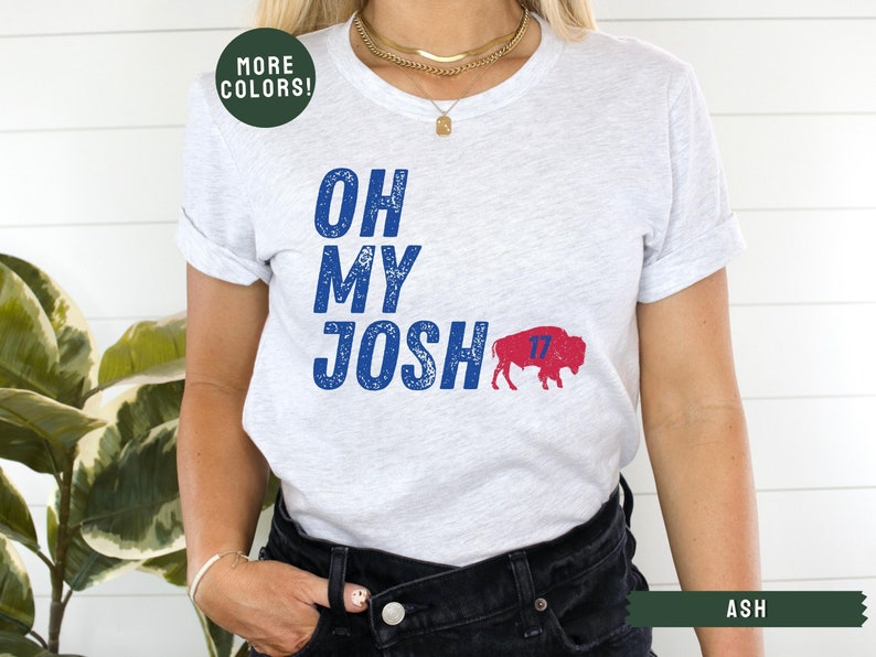 May include: A white t-shirt with blue text that reads "Oh My Josh" and a red and blue buffalo with the number 17 on it. The shirt is being worn by a person with a gold necklace.