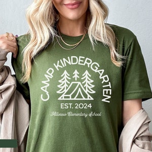 May include: Olive green t-shirt with a white graphic of a camping scene and the text "Camp Kindergarten Est. 2024 Hillview Elementary School".