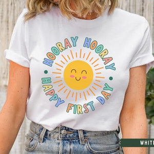 K&ouml;nnte beinhalten: Wei&szlig;es T-Shirt mit einer bunten Grafik einer l&auml;chelnden Sonne mit dem Text "Hooray Happy First Day" drumherum.