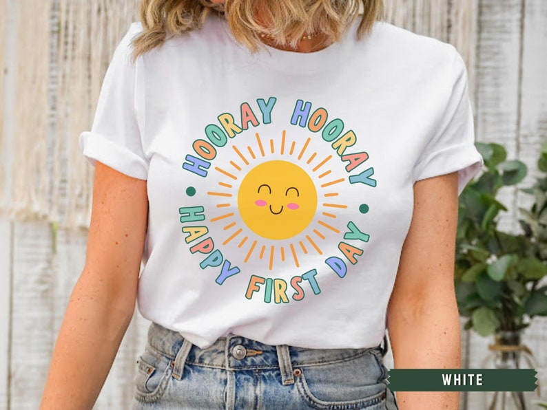 K&ouml;nnte beinhalten: Wei&szlig;es T-Shirt mit einer bunten Grafik einer l&auml;chelnden Sonne mit dem Text "Hooray Happy First Day" drumherum.