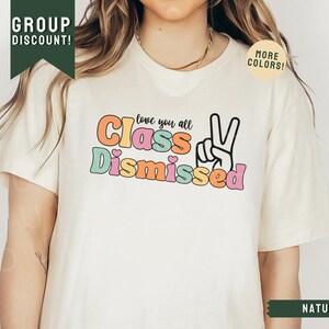 May include: A white t-shirt with the text "love you all Class Dismissed" in colorful letters with a peace sign. The shirt is on a model with long brown hair.