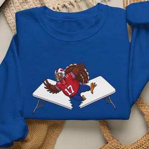 May include: A royal blue long-sleeve sweatshirt featuring a graphic of a brown turkey wearing a red football jersey with the number 17 and a white helmet, shown jumping through a white folding table that is broken into two separate pieces.