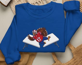 May include: A royal blue long-sleeve sweatshirt featuring a graphic of a brown turkey wearing a red football jersey with the number 17 and a white helmet, shown jumping through a white folding table that is broken into two separate pieces.
