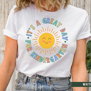 May include: White t-shirt with a colorful graphic that says "It's a Great Day for Kindergarten" with a smiling sun in the center.