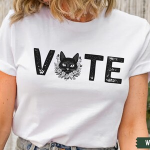 Puede incluir: Camiseta blanca con la palabra "VOTE" impresa en negro con un gr&aacute;fico de gato negro en el centro de la palabra.