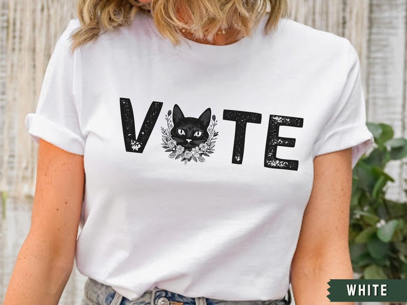 Puede incluir: Camiseta blanca con la palabra "VOTE" impresa en negro con un gr&aacute;fico de gato negro en el centro de la palabra.