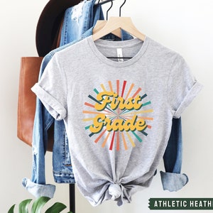 May include: A gray t-shirt with a retro-style graphic that says "First Grade" in yellow letters with a colorful sunburst design behind it. The shirt is on a hanger and has the text "Athletic Heather" at the bottom.