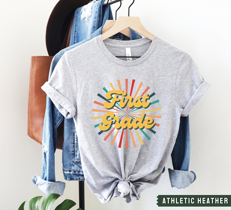 May include: A gray t-shirt with a retro-style graphic that says "First Grade" in yellow letters with a colorful sunburst design behind it. The shirt is on a hanger and has the text "Athletic Heather" at the bottom.
