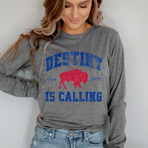 May include: A gray long-sleeve shirt with the text "DESTINY IS CALLING" in blue, and a red buffalo graphic. The shirt also includes the text "since 1960". The shirt is made of a soft, comfortable material.
