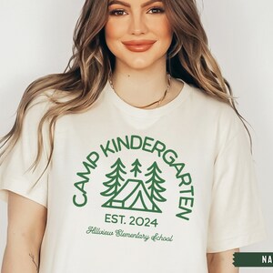 May include: A white t-shirt with a green graphic design that says "Camp Kindergarten Est. 2024 Hillview Elementary School". The graphic features a tent and pine trees.
