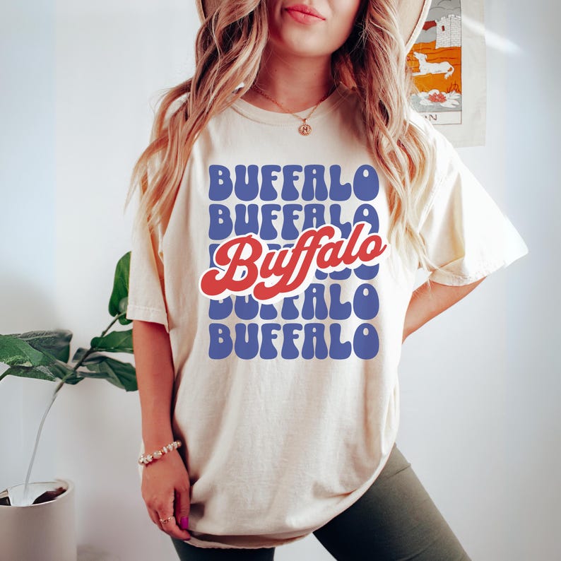 May include: A white t-shirt with a retro-style graphic design featuring the word "Buffalo" repeated in a blue and red color scheme.