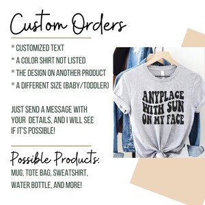 May include: A gray t-shirt with the text "ANYPLACE WITH SUN ON MY FACE" in black. The text is in a retro font style. The t-shirt is on a hanger and is displayed against a denim jacket background.