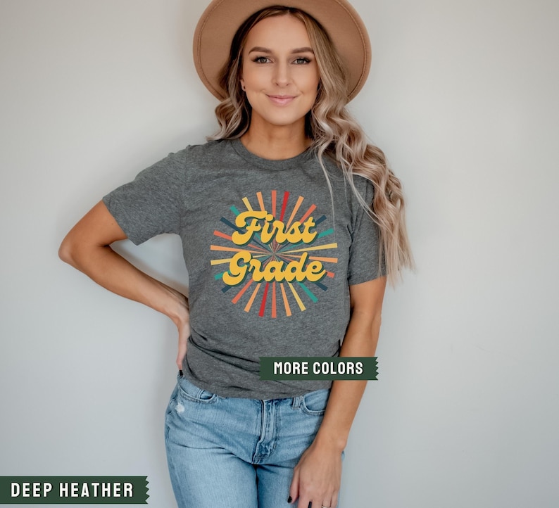 May include: A heather grey t-shirt with a retro-style graphic that says "First Grade" in yellow letters with a colorful sunburst design behind it.