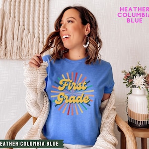 May include: A heather columbia blue t-shirt with a retro-style graphic that says "First Grade" in yellow letters with a rainbow-colored sunburst behind it.