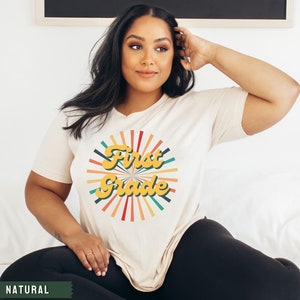May include: A white t-shirt with a retro-style graphic that says "First Grade" in yellow letters with a colorful sunburst design behind the text.