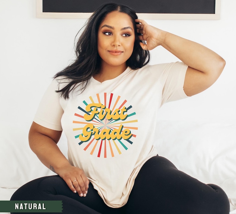 May include: A white t-shirt with a retro-style graphic that says "First Grade" in yellow letters with a colorful sunburst design behind the text.