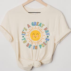 Peut inclure: Un t-shirt de couleur naturelle avec un motif de soleil jaune souriant entour&eacute; du texte "IT'S A GREAT DAY TO BE IN PRE-K" en lettres color&eacute;es et arrondies. Le motif est centr&eacute; sur la poitrine du t-shirt, qui est pr&eacute;sent&eacute; sur un cintre.