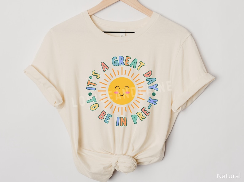 Peut inclure: Un t-shirt de couleur naturelle avec un motif de soleil jaune souriant entour&eacute; du texte "IT'S A GREAT DAY TO BE IN PRE-K" en lettres color&eacute;es et arrondies. Le motif est centr&eacute; sur la poitrine du t-shirt, qui est pr&eacute;sent&eacute; sur un cintre.