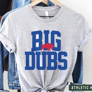 May include: A gray t-shirt with the words "BIG DUBS" in blue letters and a red buffalo silhouette. The shirt is labeled "ATHLETIC HEATHER".
