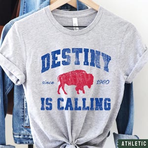May include: Gray t-shirt with a blue and red vintage graphic that says "Destiny since 1960 is calling". The graphic features a red buffalo silhouette.