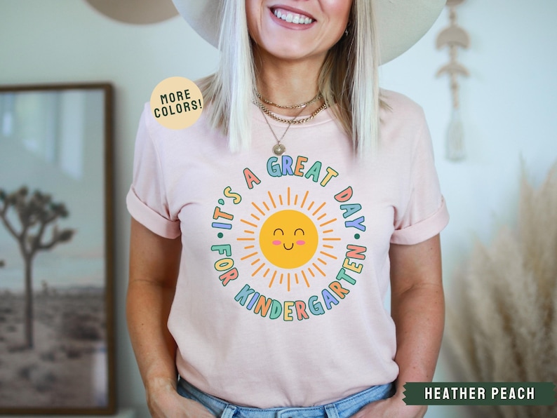 May include: A pink t-shirt with a colorful graphic of a smiling sun and the text "It's a Great Day for Kindergarten".