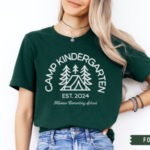 May include: A dark green t-shirt with a white graphic of a tent and trees. The text "Camp Kindergarten Est. 2024 Hillview Elementary School" is printed on the shirt.