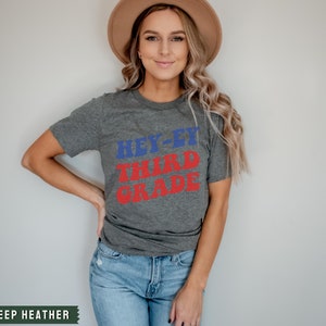 May include: A grey t-shirt with the text "Hey-ey Third Grade" in red and blue letters. The shirt is being worn by a person with long blonde hair and a brown hat.