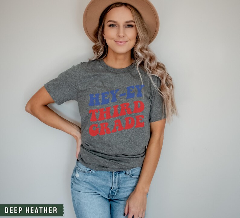 May include: A grey t-shirt with the text "Hey-ey Third Grade" in red and blue letters. The shirt is being worn by a person with long blonde hair and a brown hat.