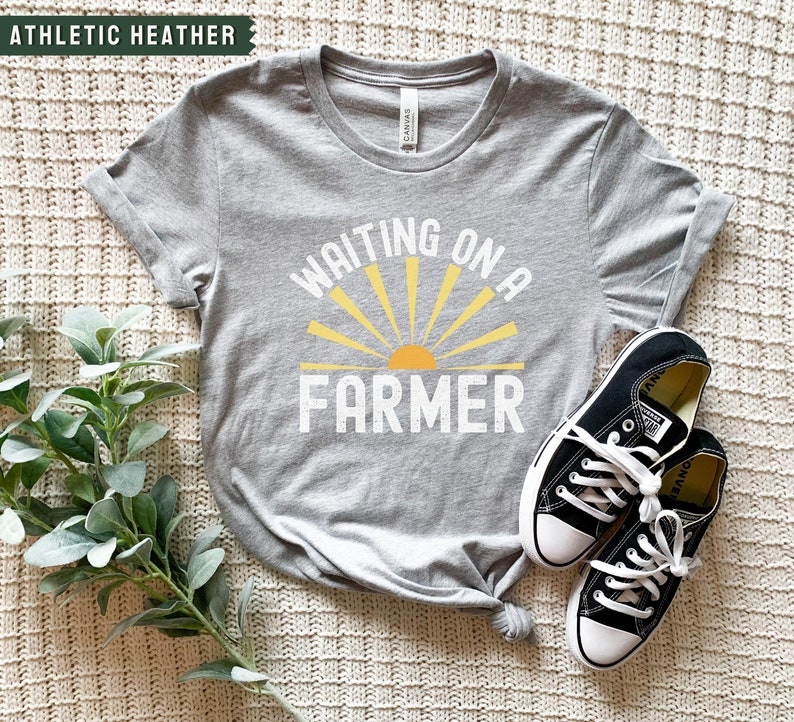 Może przedstawiać: Szary T-shirt z napisem "Waiting on a Farmer" w kolorze białym i ż&oacute;łtym graficznym słońcem.