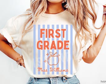 May include: Ivory-colored t-shirt with a graphic for First Grade. The design features vertical blue and white stripes, with the words "FIRST GRADE" in orange. An orange apple graphic is below the words, with the name "Miss Williams" in cursive.